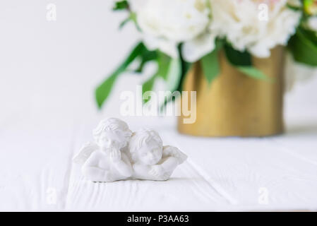 Vicino la statuetta di due antichi piccoli angeli incantevole del gesso sul bianco tavolo in legno con bouquet di peonie bianco nella ciotola vintage. Amore bac Foto Stock