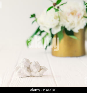 Vicino la statuetta di due antichi piccoli angeli incantevole del gesso sul bianco tavolo in legno con bouquet di peonie bianco nella ciotola vintage. Amore bac Foto Stock