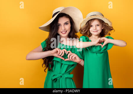 Bella madre in piedi con la bambina nella stessa green vestiti, cappelli di paglia tenendo le mani in forma di cuore e guardando la fotocamera su sfondo giallo Foto Stock