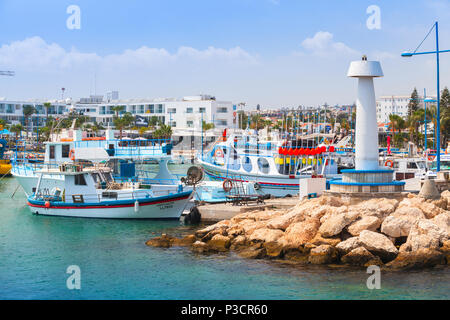 Ayia Napa, Cipro - 12 Giugno 2018: Agia Napa Marina vista con ormeggiate barche da pesca e bianco Torre Faro sulla scogliera, i turisti a piedi sulla costa Foto Stock