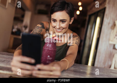 Donna sorridente prendendo un selfie con un smoothie collocato di fronte a lei utilizzando un telefono cellulare per il suo cibo blog. Blogger alimentare a scattare foto per il suo blog Foto Stock