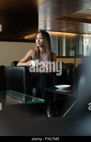 Asian imprenditrice guardando lontano tenendo tazza da caffè e telefono cellulare Foto Stock
