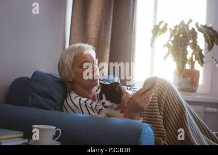 Senior donna seduta su un divano con il suo gatto mentre si utilizza il telefono cellulare nel soggiorno Foto Stock