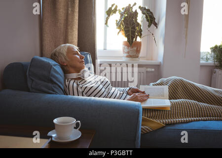 Senior donna prendendo un pisolino sul divano mentre la lettura del libro in salotto Foto Stock