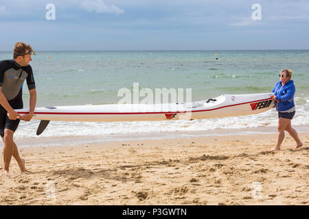 L uomo e la donna che la porta di un surf ski surfski lungo il litorale di Branksome Chine, Poole, Dorset, England Regno Unito nel mese di giugno Foto Stock