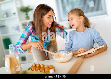 La madre e la bambina di miscelazione di pasta nella cucina domestica e la figlia sta leggendo la ricetta dal libro di cucina. Madre impara la sua figlia per rendere la cooki Foto Stock