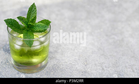 Un bicchiere di verde chiaro liquido viene raffreddato con cubetti di ghiaccio e appannato. Si erge su una concreta della metropolitana. Le foglie di menta dell'erba " Marocco " Menta decorat Foto Stock