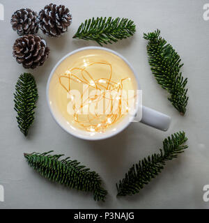 Vista dall'alto di una tazza da bianco a caldo con le luci di Natale all'interno circondato da pigne e rami su un tavolo bianco. Concetto di natale. Formato quadrato. Foto Stock