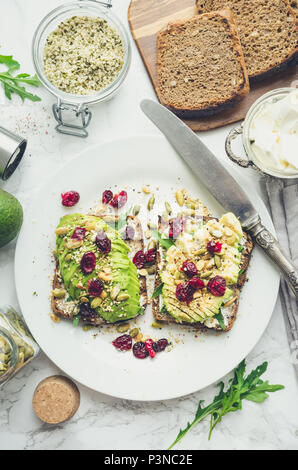 Una sana avocado toast per la prima colazione o il pranzo con pane di segale, crema di formaggio, rucola, tagliata a fette di avocado, essiccato cranberry, zucca, di canapa e di semi di sesamo. Foto Stock