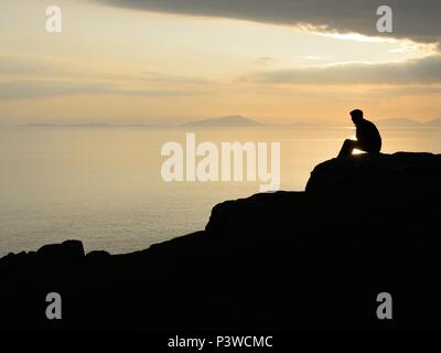 A profilarsi l'uomo guarda al mare al tramonto da una scogliera a Neist punto sull'Isola di Skye in Scozia, Regno Unito Foto Stock