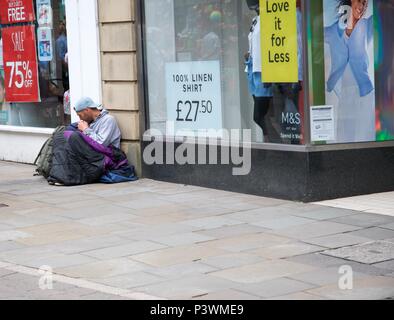 Un uomo apparentemente senzatetto, siede sul terreno al di fuori di un negozio a Buxton, High Peak, Derbyshire, Regno Unito Foto Stock