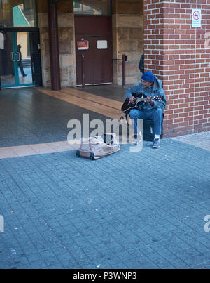 Un busker seduto su un altoparlante quaresima contro un muro di mattoni di suonare una chitarra con una borsa di fronte a lui in attesa di una donazione, Sydney, Australia Foto Stock