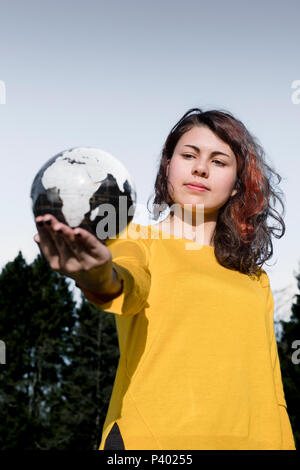 Il mondo intero nella sua mano: la giovane donna in eleganti maglione giallo tenendo la terra nella sua mano nella parte anteriore del cielo e alberi. Salvare il concetto di massa. Foto Stock