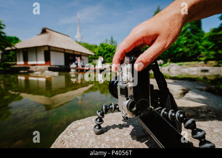 Fotografia maschio di manipolazione a mano un vecchio vintage fotocamera su treppiede nella parte anteriore del laghetto di acqua con un punto di riferimento della casa giapponese e coppie in viaggio di nozze nel parco Planten un Blomen Park Hamburg, Germania Foto Stock