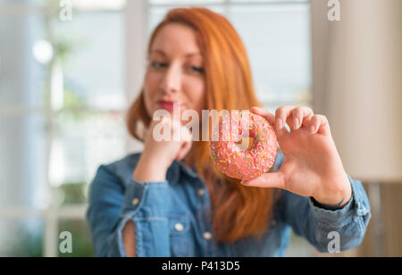 Redhead donna ciambella di contenimento a casa faccia seriamente pensando di domanda, molto confusa idea Foto Stock