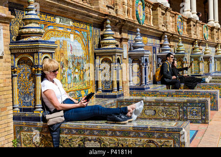 Donna di relax al sole durante la lettura da un tablet su uno della provincia piastrellato alcove, Plaza de Espana, Andalusia, Spagna. Foto Stock