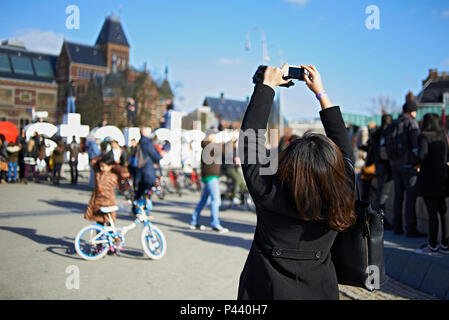 Oltre la spalla colpo di un turista femminile nella folla di scattare una foto utilizzando un telefono cellulare nel centro della città di Amsterdam in inverno il sole Foto Stock