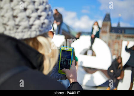 Oltre la spalla colpo di un turista femminile nella folla di scattare una foto utilizzando un telefono cellulare nel centro della città di Amsterdam in inverno il sole Foto Stock