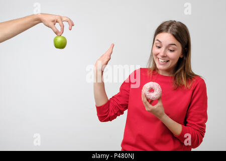 Cibo sano e concetto di dieta. Bella giovane donna scegliere tra frutta e dolci. Lei preferisce una ciambella rosa invece di mela verde Foto Stock