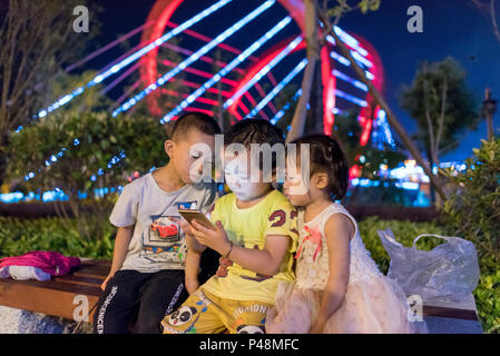 Bambini video gioco sul telefono nel parco della città vicino al neon, ponte di notte la vita di strada, Libo, Guizhou, Cina Foto Stock