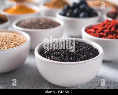 Sesamo nero in piccola ciotola bianco e altri superfoods sullo sfondo. Messa a fuoco selettiva. Superfoods diversi ingredienti. Concetto e illustrazione per la super e detox food Foto Stock