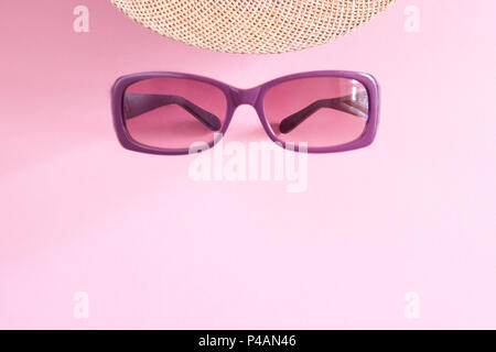 Occhiali da sole viola e parte del cappello di paglia su sfondo rosa con copia spazio. Foto Stock