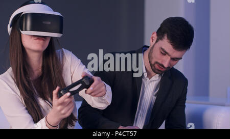 Sorridente giovane uomo con il telefono cerca di fermare la ragazza in VR auricolare dalla riproduzione di tanto Foto Stock