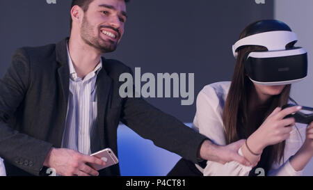 Uomo sorridente con il telefono cellulare tenta di distrarre il suo partner di sesso femminile da riproduzione di gioco di realtà virtuale Foto Stock