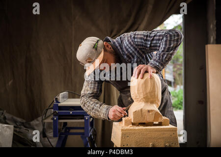 Close up di un falegname, builder in abiti da lavoro sega per tagliare fuori la scultura in legno da un uomo di testa in officina, intorno un sacco di strumenti in legno,,furn Foto Stock