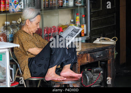 Hanoi, Vietnam - Marzo 15, 2018: Locale senior signora seduta nel suo negozio e leggendo un giornale Foto Stock