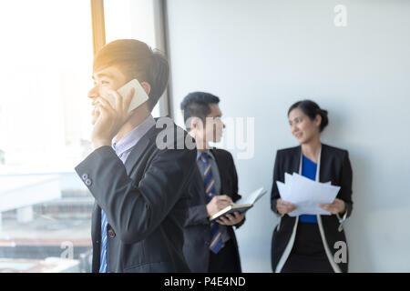 Asian business man o un avvocato di parlare tramite smartphone sorridendo felice all'interno di edificio per uffici con i suoi colleghi sullo sfondo. Foto Stock