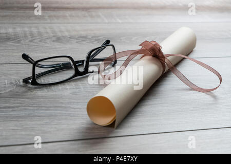 Un diploma arrotolato e legato con un nastro rosso sul tavolo con un paio di occhiali Foto Stock