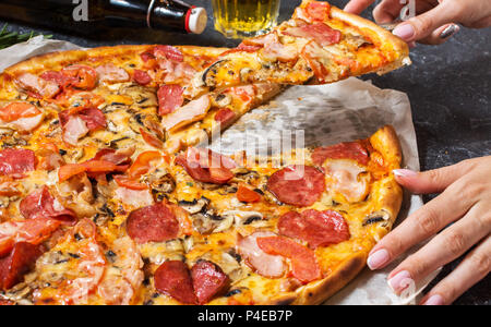 Mani femminili tenere un pezzo freschi fatti in casa la pizza con salsiccia per pizza, prosciutto, formaggio su rustico in pietra dello sfondo. Foto Stock