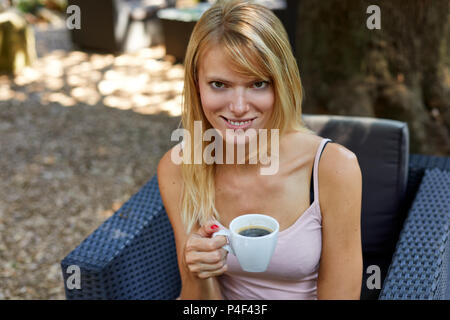 Bella giovane donna in abiti estivi seduto sulla poltrona da giardino con tazza di caffè caldo e guardando la telecamera sorridente su sfondo sfocato Foto Stock