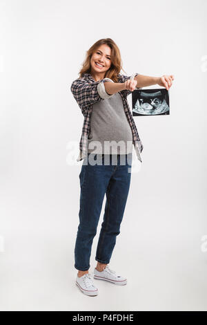 Sorridente donna incinta che mostra di scansione ad ultrasuoni in mani isolato su grigio Foto Stock