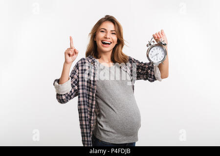 Felice donna incinta con orologio in mano rivolto verso l'alto isolato su grigio Foto Stock