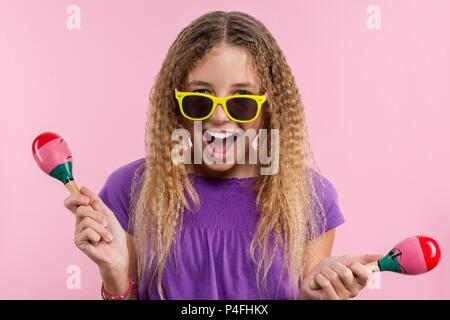 Allegro teen ragazza con lunghi biondi capelli ricci. In luminoso giallo bicchieri ella danze con le maracas su uno sfondo rosa Foto Stock