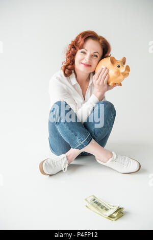 Felice donna matura con banconote di dollari e salvadanaio, isolato su bianco Foto Stock