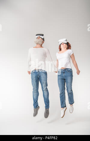 Coppia senior jumping mentre utilizzando la realtà virtuale di cuffie, isolato su bianco Foto Stock