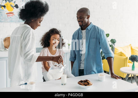 African American donna versando il latte in vetro con la famiglia vicino in cucina a casa Foto Stock