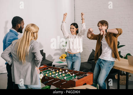 Happy business partner celebrando la vittoria nel calcio da tavolo con le braccia in alto e il pollice verso il basso i gesti di fronte ai colleghi Foto Stock