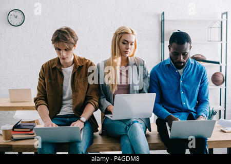 Tre multiculturale di colleghi di lavoro che utilizzano portatili e seduta sul tavolo in ufficio moderno Foto Stock