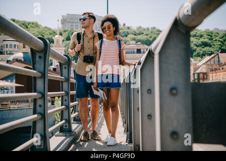 Interracial paio di giovani viaggiatori con fotocamera e zaini camminando sul ponte Foto Stock