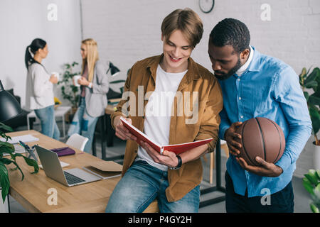 Imprenditore sorridente che mostra un libro di testo di African partner americano tenendo palla, due femmina i colleghi in piedi dietro con tazze da caffè Foto Stock