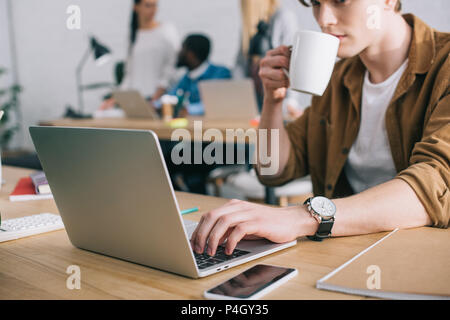 Ritagliato shot del giovane imprenditore a bere caffè e utilizzando il portatile in tabella e colleghi lavorando dietro Foto Stock