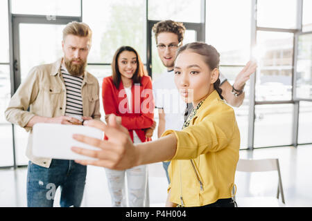 Vista ritagliata di smartphone in mano sulla donna prendendo selfie con diversi business team di lavoro sul progetto in area di lavoro di luce Foto Stock