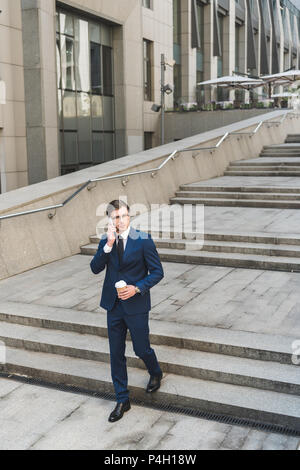 Bel giovane imprenditore nel vestito elegante con caffè per andare a parlare al telefono mentre andando giù per le scale Foto Stock