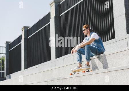 Elegante giovane uomo in denim vestiti seduti sulle scale con longboard guardando lontano Foto Stock