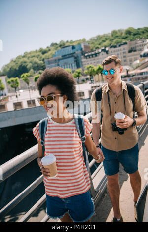 Giovane coppia multiculturale di turisti con tazzine di caffè e fotocamera camminando sul ponte Foto Stock