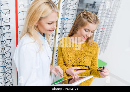 Sorridente oculista con tavoletta digitale consentendo al cliente di scegliere gli occhiali in ottica Foto Stock